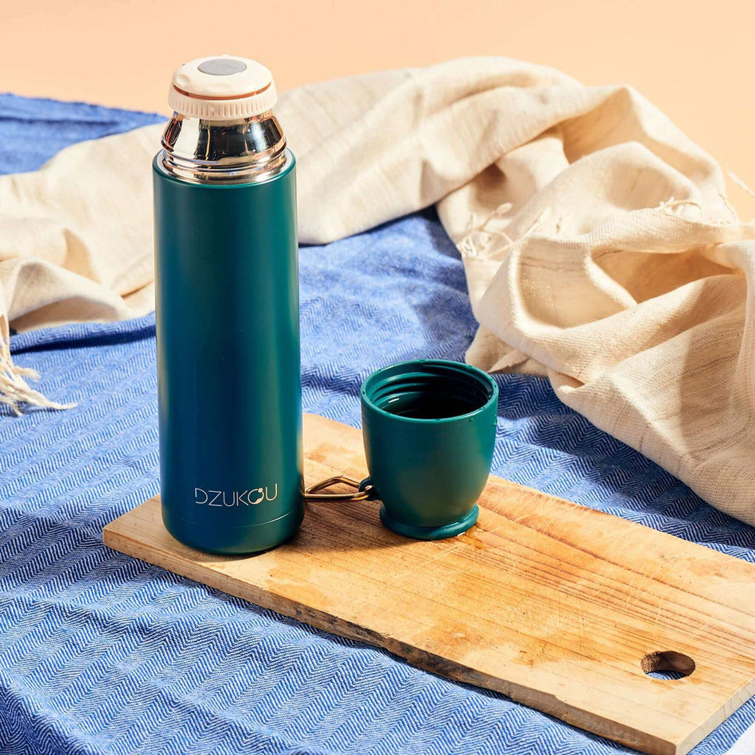 A stainless steel thermos flask and a cup placed on a wooden board with a eri silk stole in a background, suggesting a outdoor beverage setting.