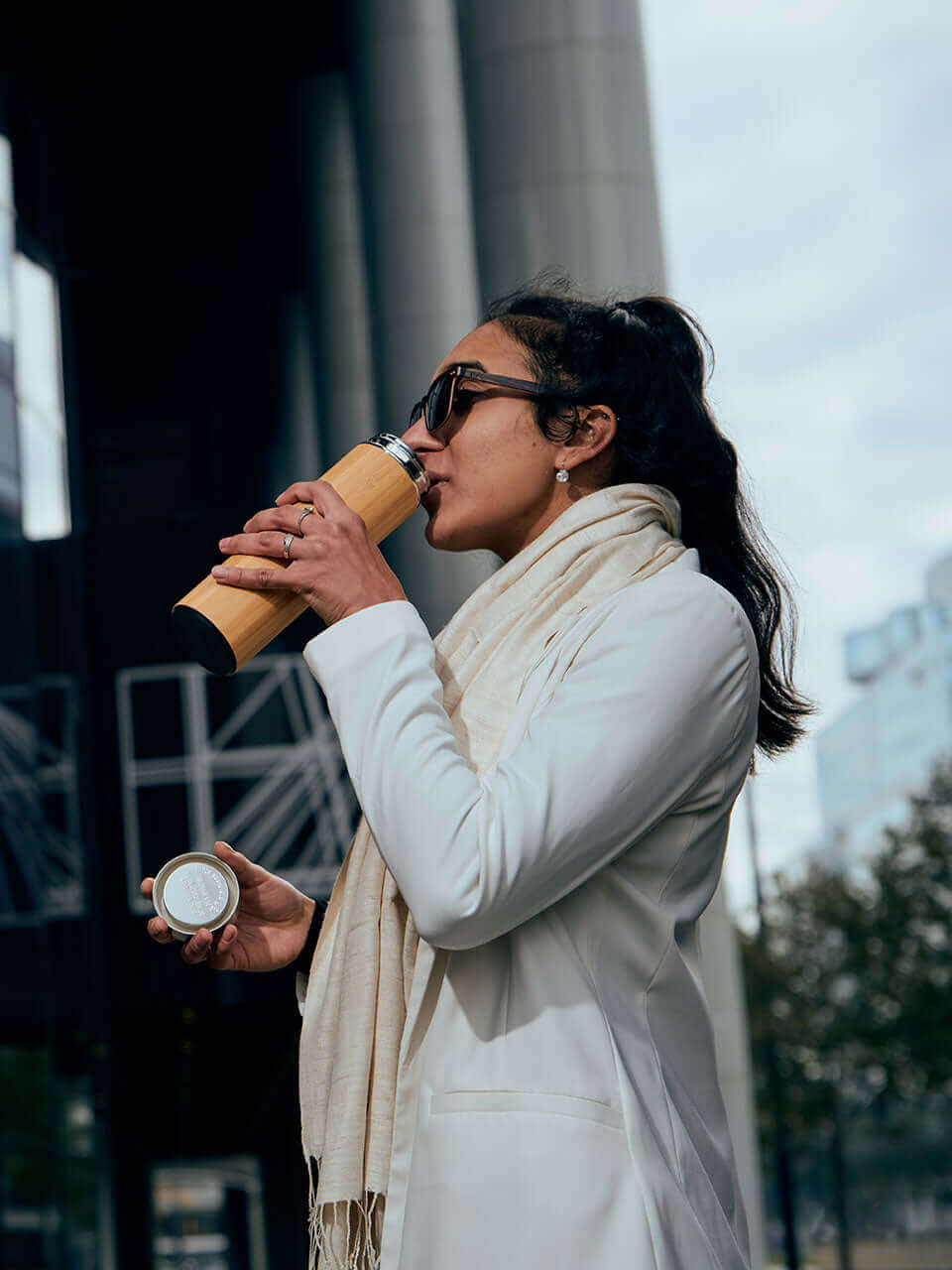 A woman in a white coat enjoys a cup of coffee from a bamboo thermos bottle, exuding a sense of warmth and comfort.