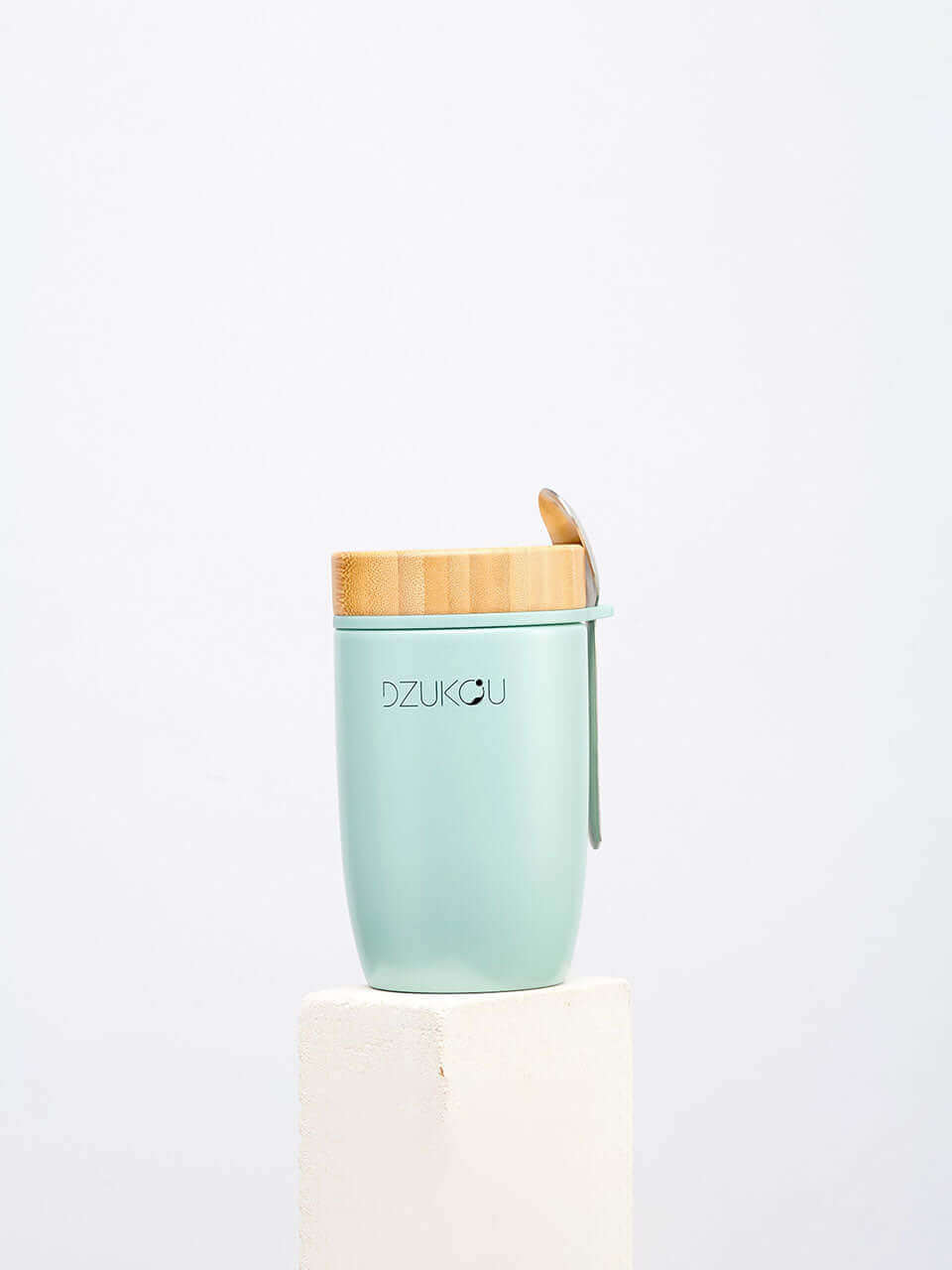 A bamboo green stainless steel lunchpot with a wooden lid and a spoon placed on a white block, showcasing a simple and elegant design.