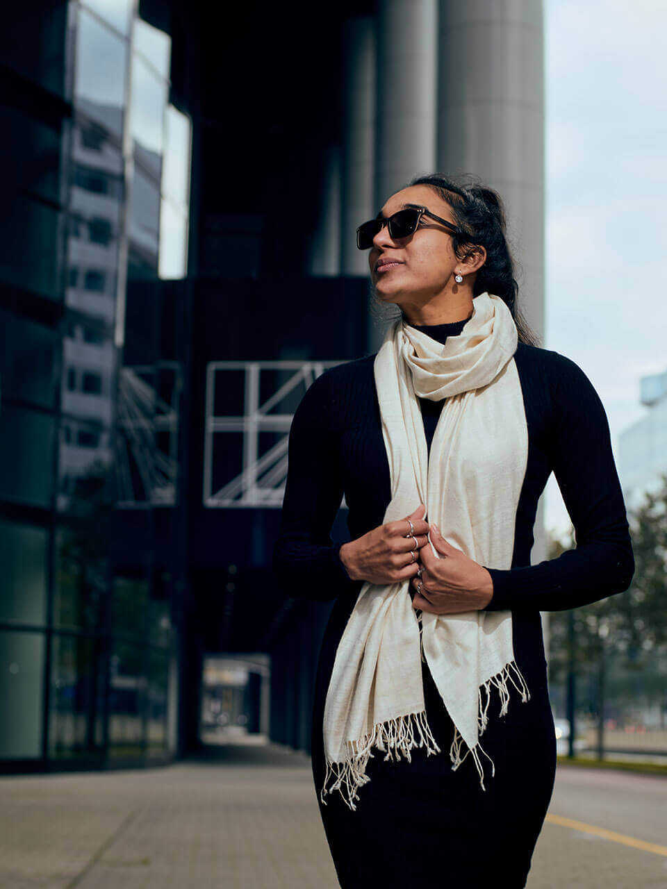 A woman in a black dress and scarf stands elegantly in front of a building, showcasing a poised and stylish demeanor.