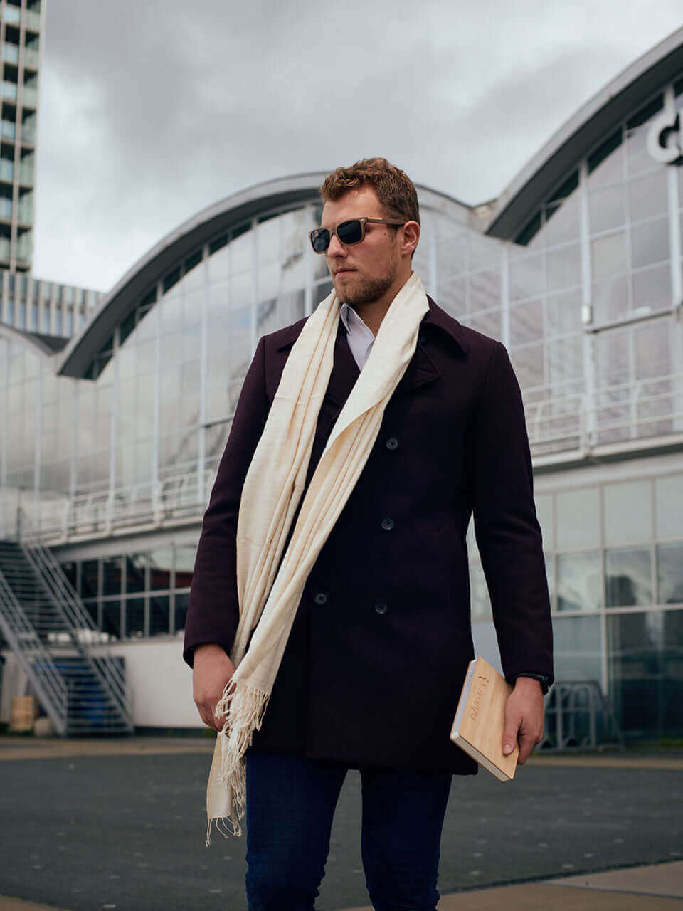 A man wearing a wooden sunglasses along with a coat and scarf strolls through an urban setting, surrounded by city buildings and streets.